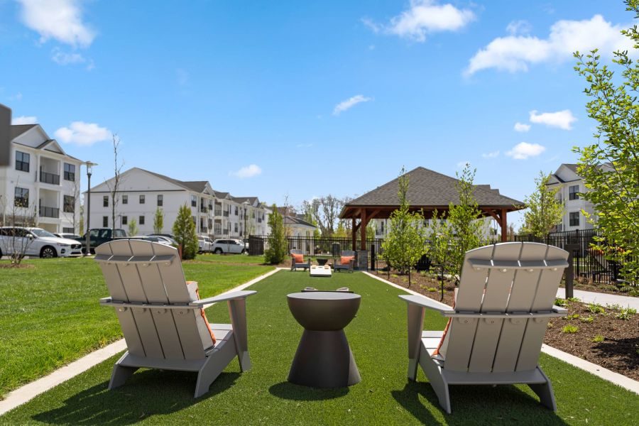 Two Adirondack chairs and a fire pit on artificial grass, with apartments and a gazebo in the background.