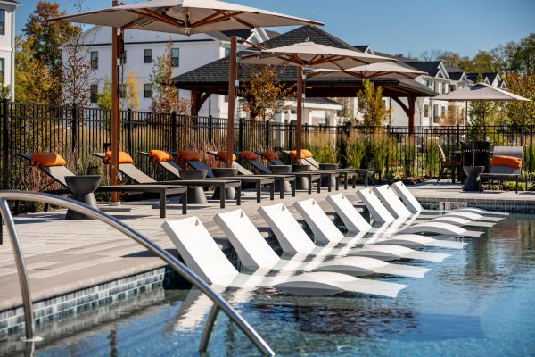 Poolside loungers partially in water, sun umbrellas, and orange-cushioned chairs beside a fenced garden area.
