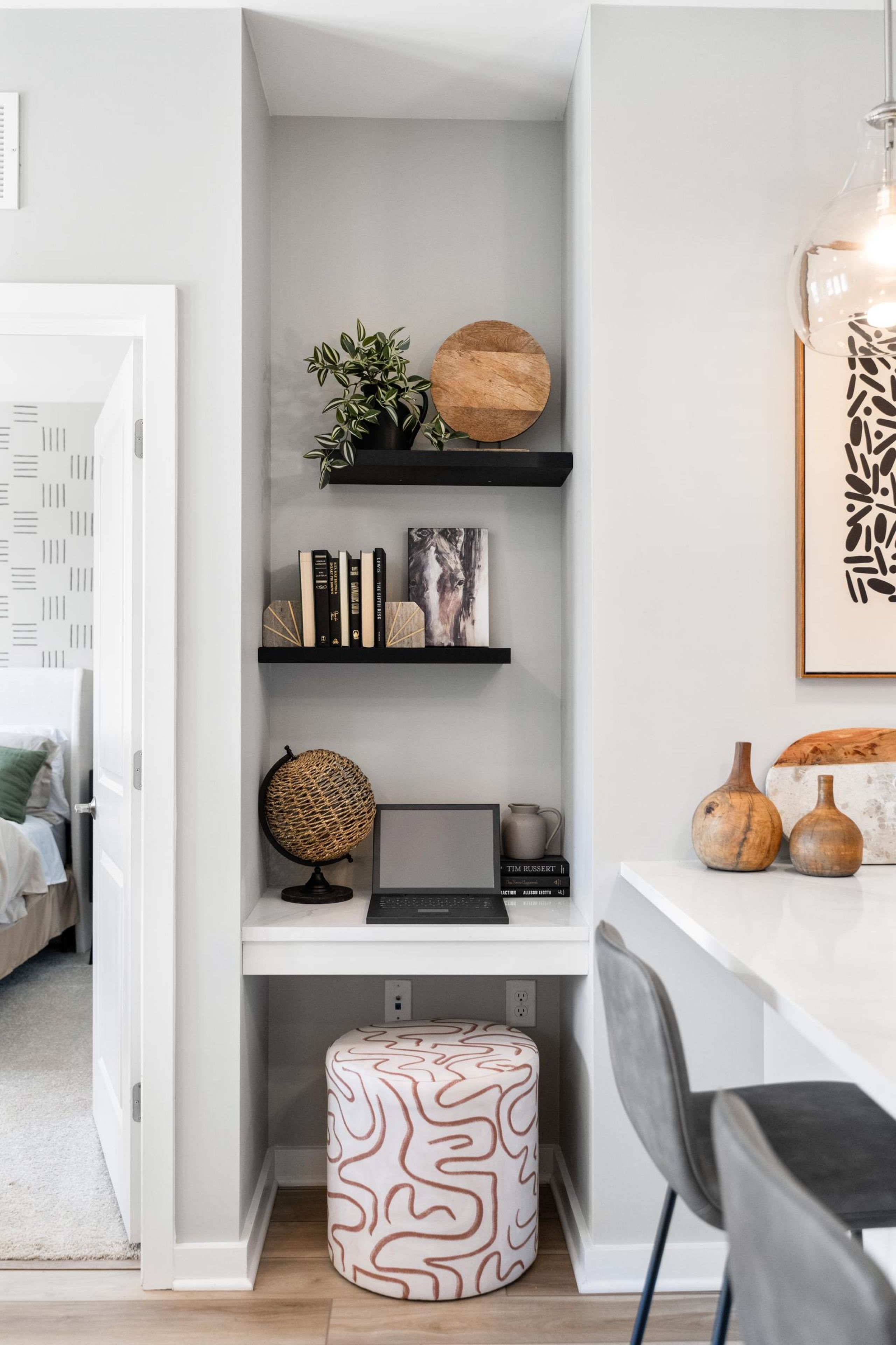 Small home office nook with shelves, decor, a laptop, globe, and a patterned ottoman under a white desk.
