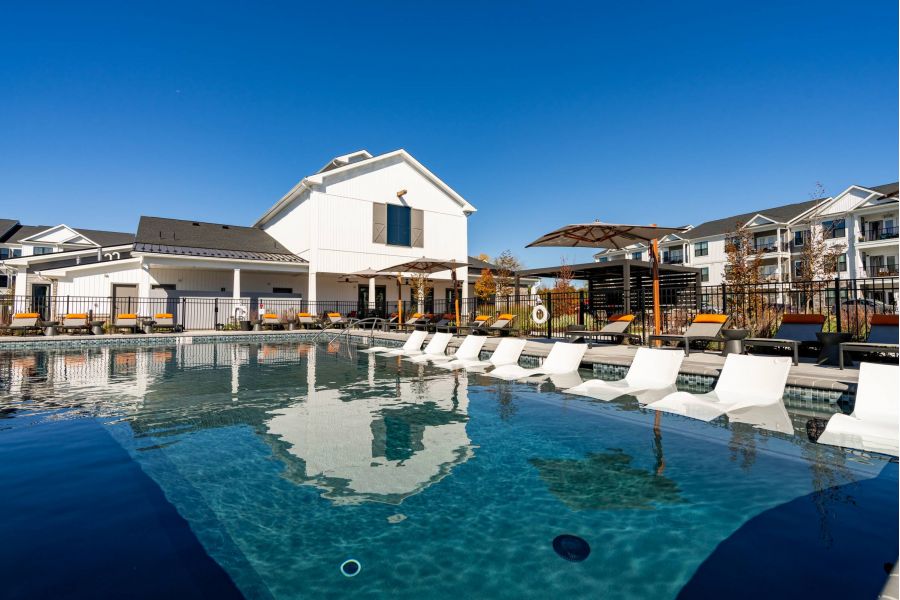 Outdoor pool with in-water lounge chairs, sun umbrellas, and modern apartments in the background under a clear sky.