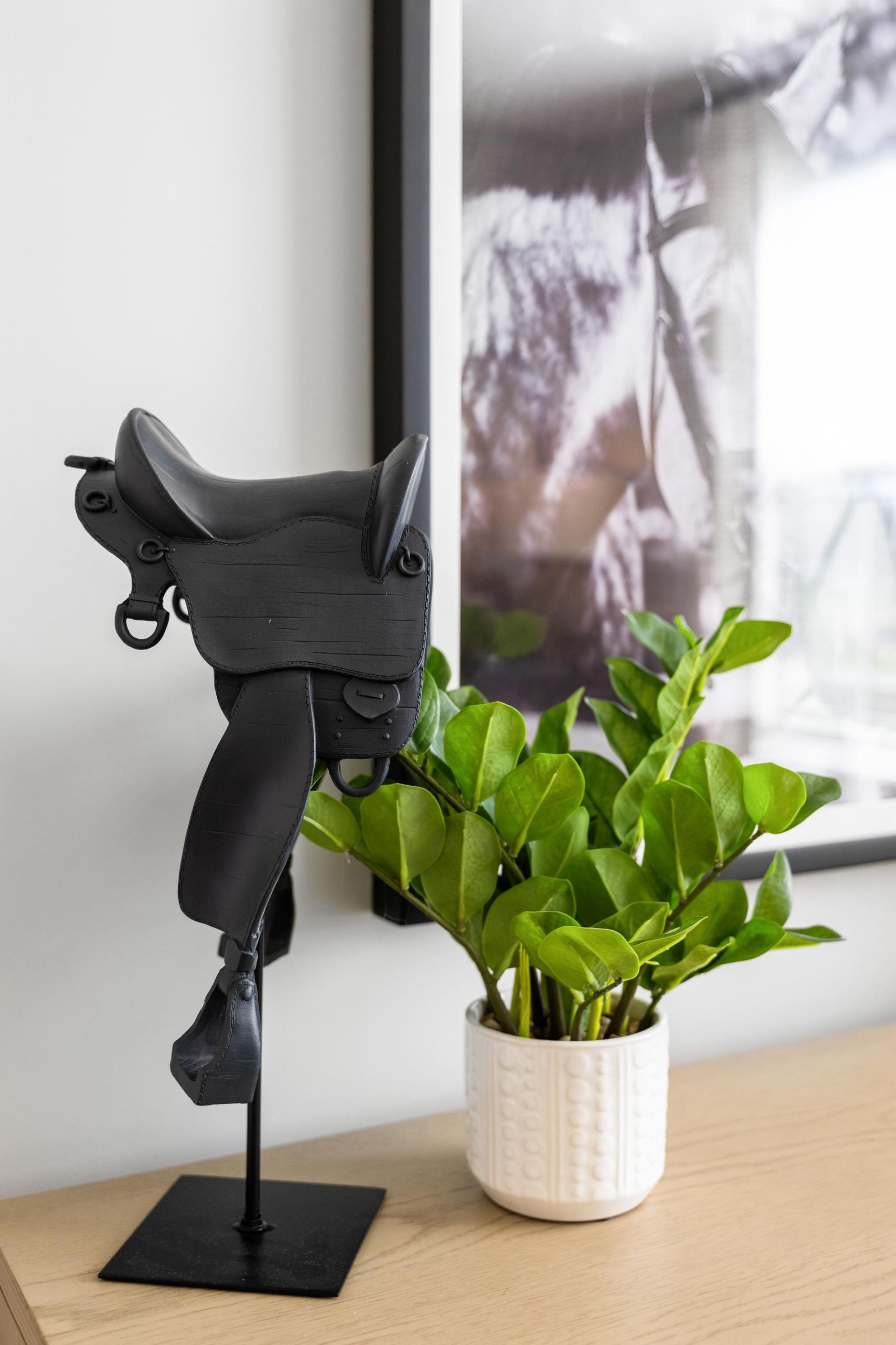 A small black saddle model on a stand beside a potted green plant on a wooden surface.