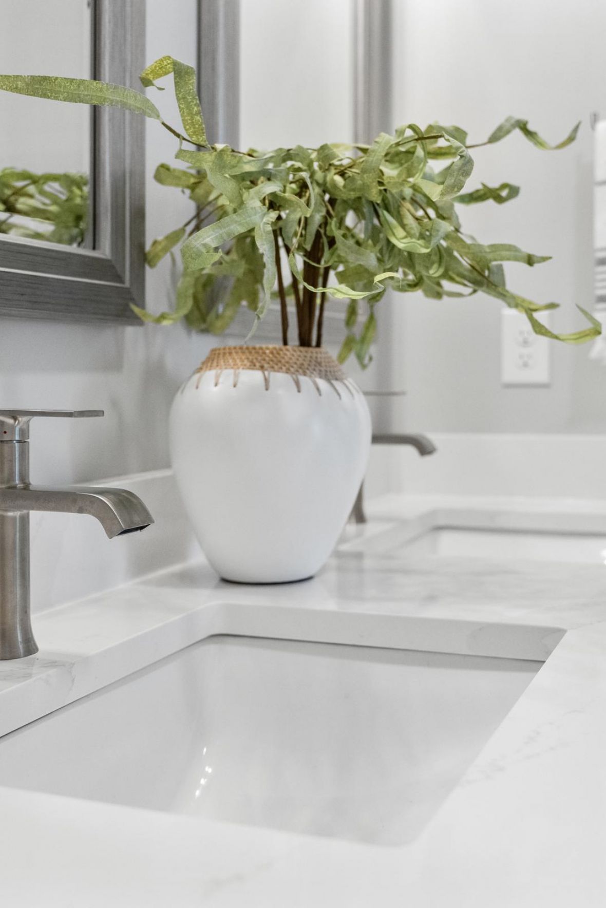 Modern white bathroom with double sinks, silver faucets, a potted plant, and a towel hanging on the wall.