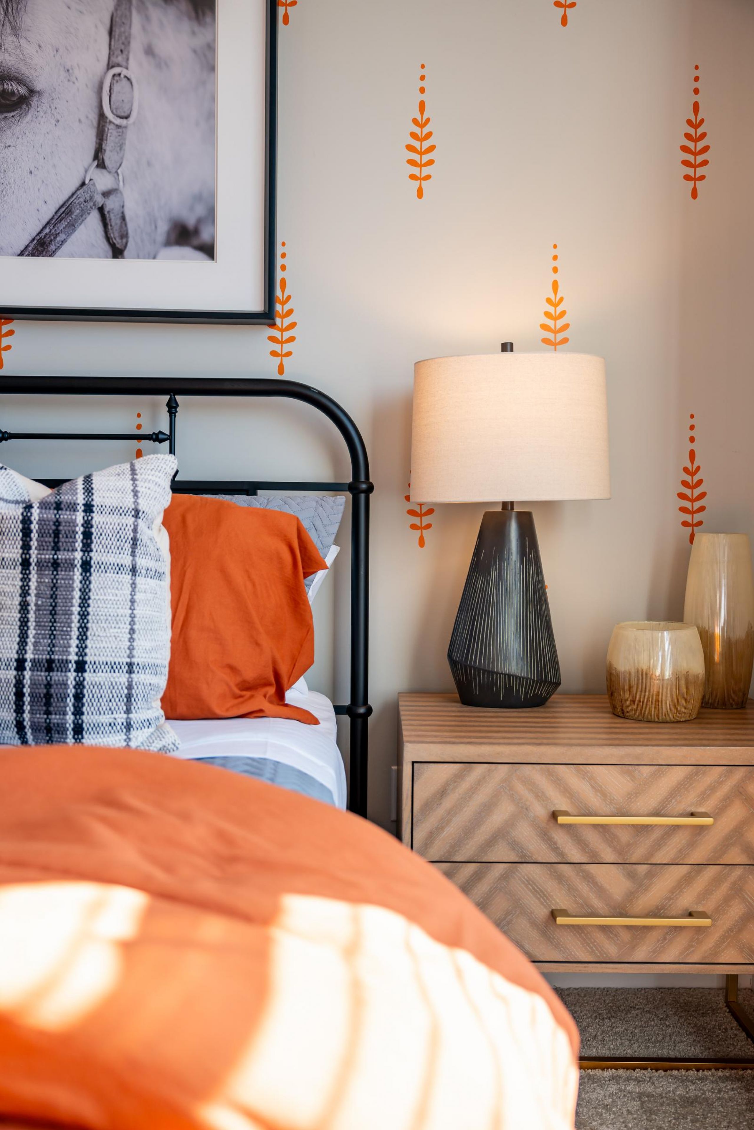 A cozy bedroom with orange bedding, plaid pillow, lamp, and decorative vases on a wooden nightstand.