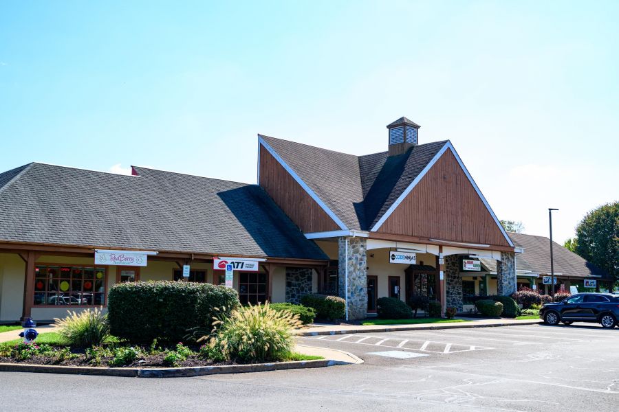 A small shopping plaza with stone accents, storefronts, and a parking lot on a sunny day.