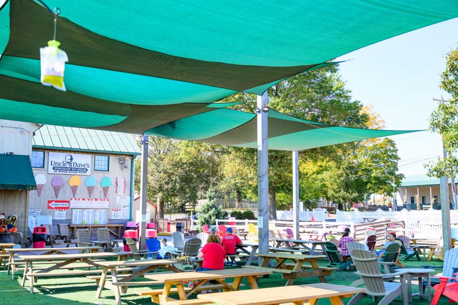 Outdoor picnic area with green shade sails, people sitting at tables, and an ice cream stand in the background.