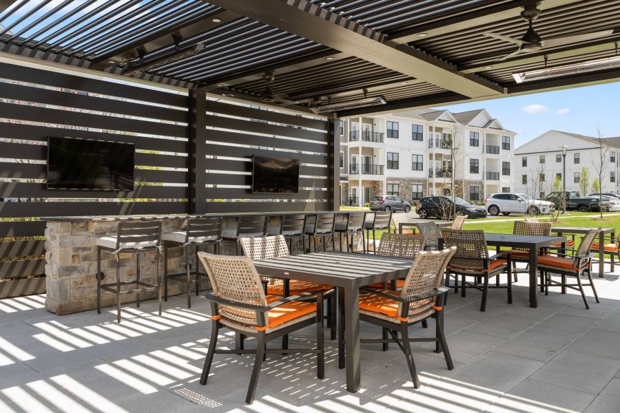 Outdoor patio with tables, chairs, bar seating, TVs, and modern apartments in the background on a sunny day.