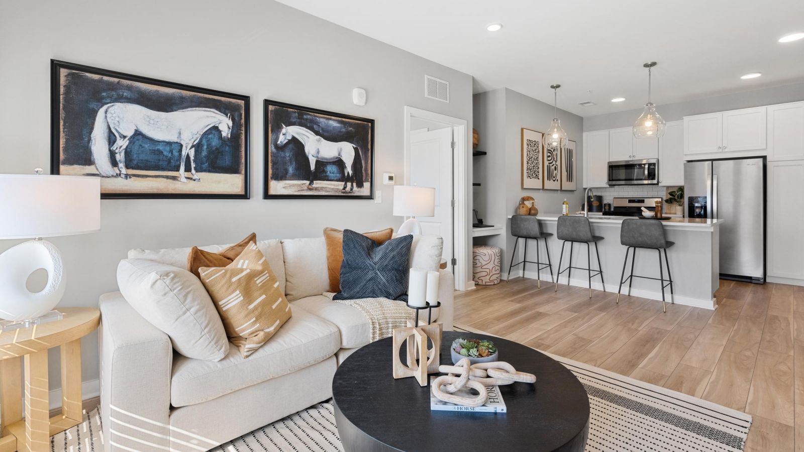 Modern living room with a white sofa, horse artwork, open kitchen, and gray barstools at the counter.