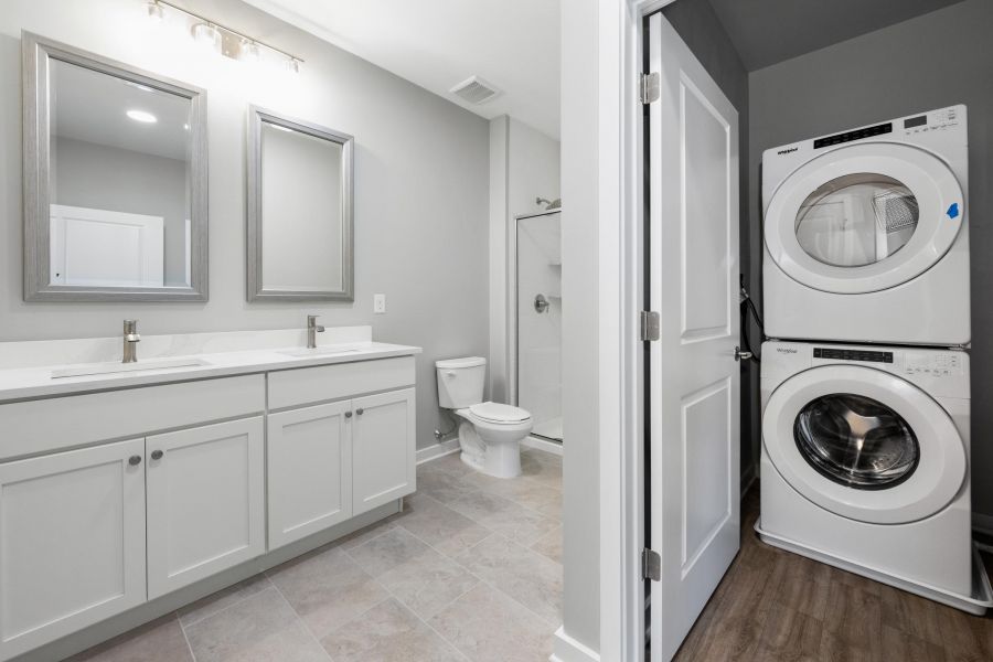 Modern bathroom with double sinks, a toilet, shower, and stacked washer and dryer in a laundry nook.