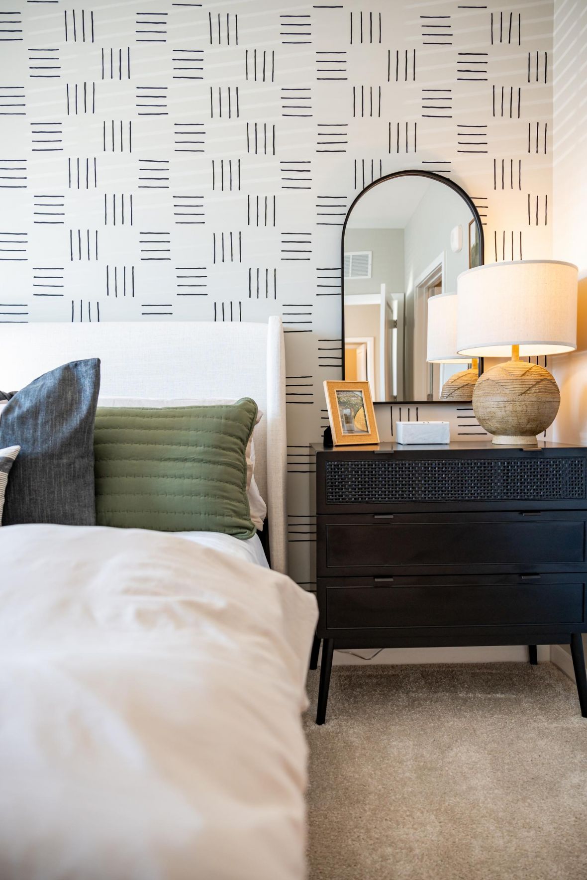 Modern bedroom with patterned wallpaper, a bed, green pillow, black nightstand, lamp, and arched mirror.