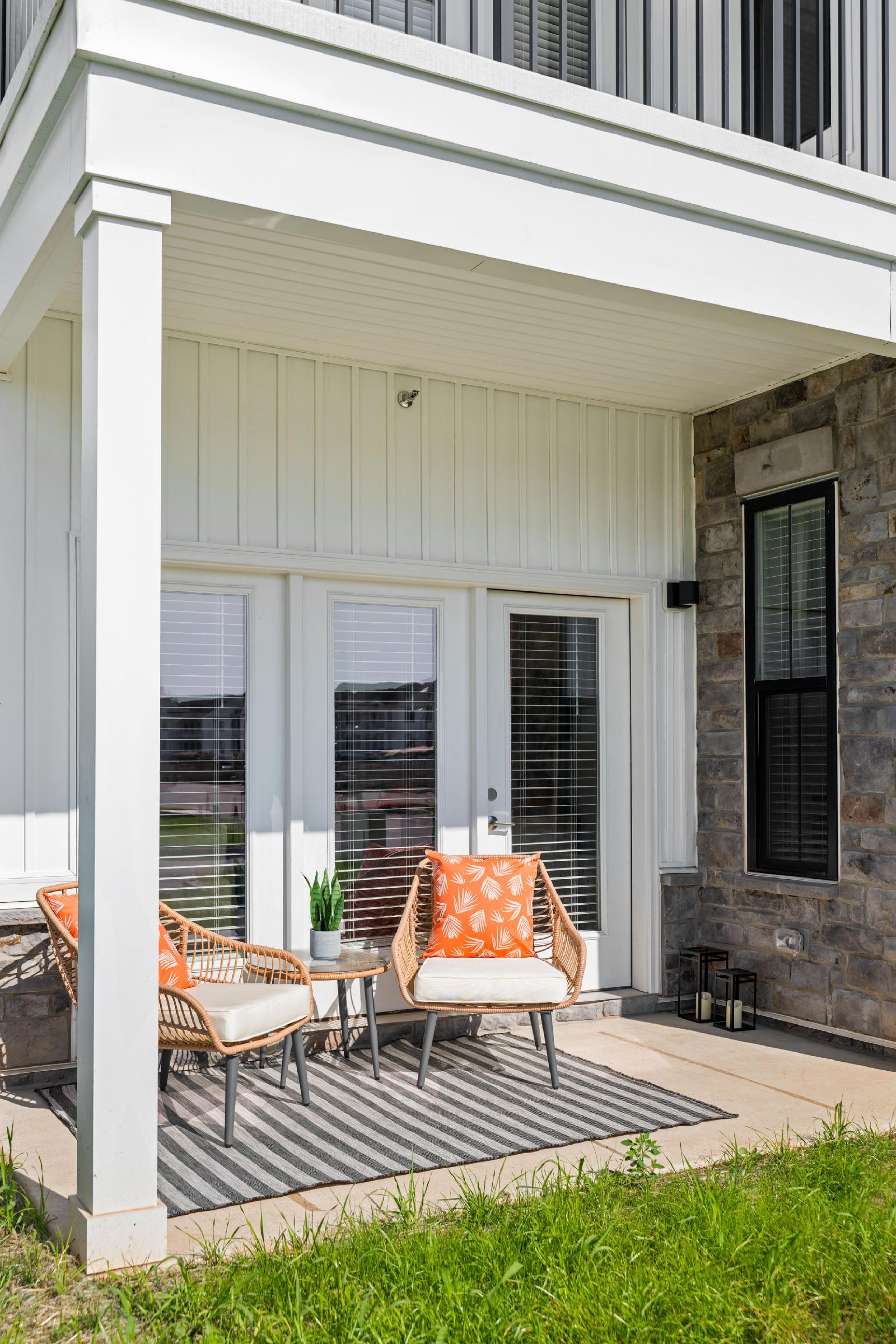 Two patio chairs with orange pillows and a small table on a striped rug outside a modern house.