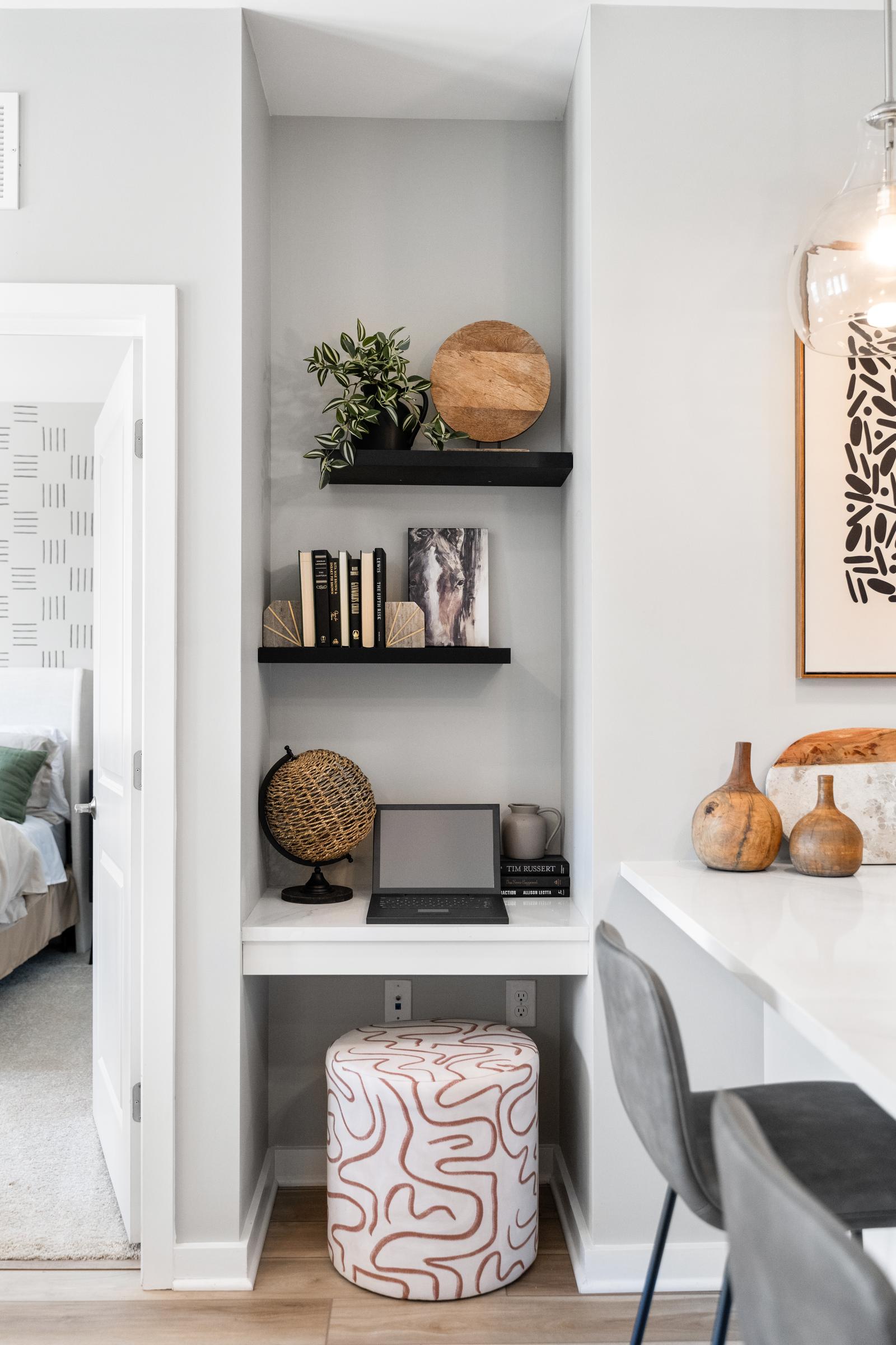Small home office nook with shelves, decor, a laptop, globe, and a patterned ottoman under a white desk.