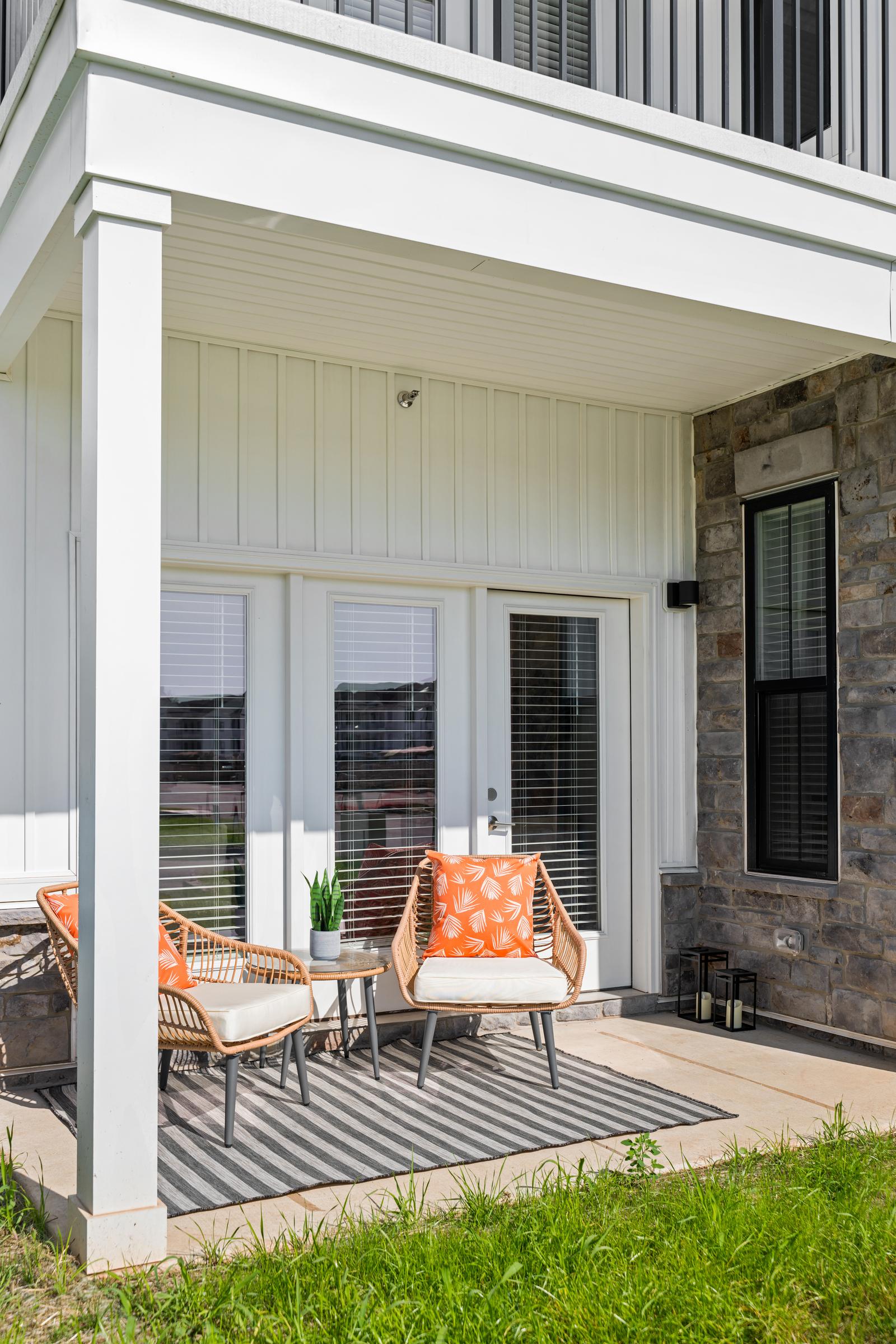 Two patio chairs with orange pillows and a small table on a striped rug outside a modern house.