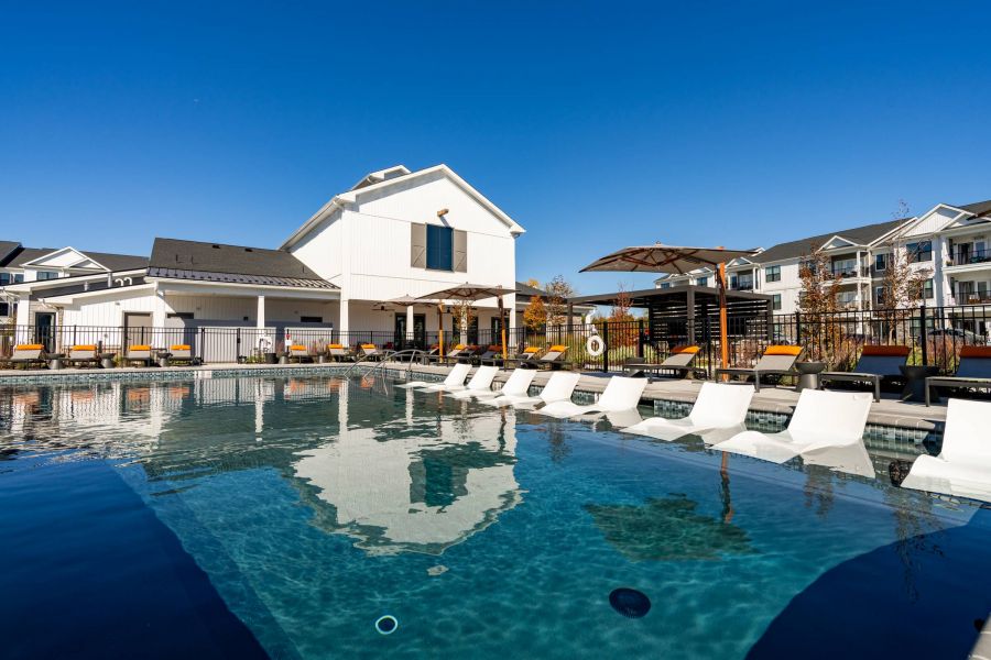Outdoor pool with in-water lounge chairs, sun umbrellas, and modern apartments in the background under a clear sky.