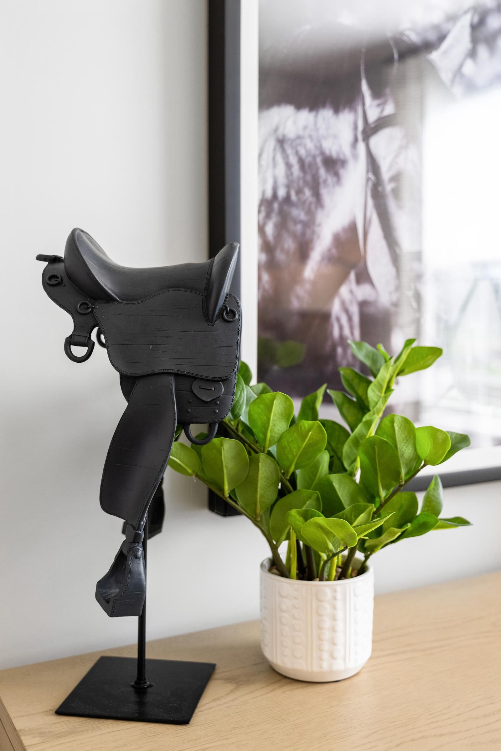 A small black saddle model on a stand beside a potted green plant on a wooden surface.