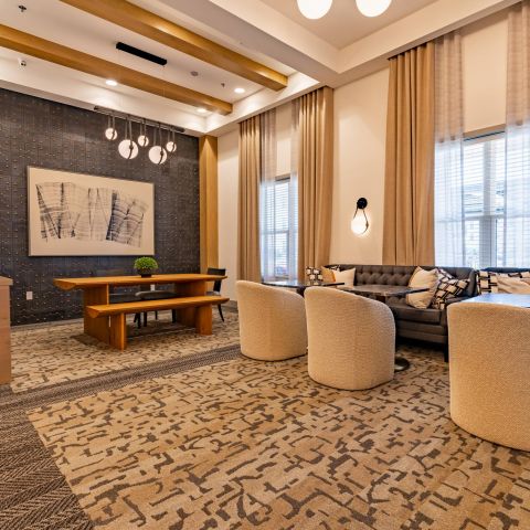 Modern lounge with a wooden table, beige chairs, tufted sofa, abstract art, and large windows with curtains.