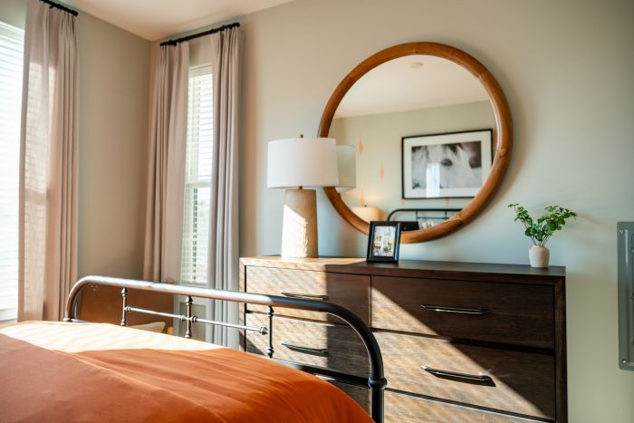 A sunlit bedroom with a dresser, round mirror, lamp, framed photo, and potted plant.